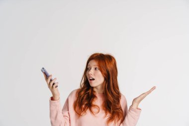 Ginger güzel, şok olmuş bir kız cep telefonu tutuyor ve beyaz arka planda el ele tutuşuyor.