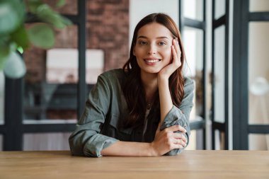 Güzel, mutlu, hoş bir kadın masada otururken kameraya gülümsüyor.