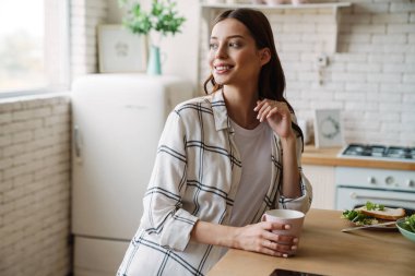 Güzel, gülümseyen bir kadın, sıcak bir mutfakta kahvaltı ederken kahve içiyor.