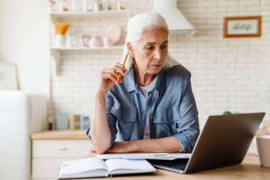 Düşünceli orta yaşlı kadın evde masada otururken bilgisayar başında çalışıyor.