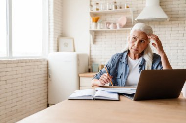 Düşünceli orta yaşlı kadın evde masada otururken bilgisayar başında çalışıyor.