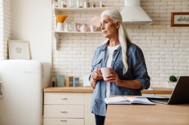 Üst düzey düşünceli kadın mutfakta dizüstü bilgisayarıyla çalışıyor, elinde bir fincan çayla.