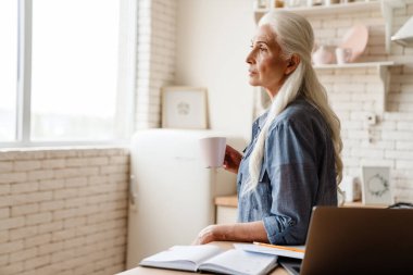 Üst düzey düşünceli kadın mutfakta dizüstü bilgisayarıyla çalışıyor, elinde bir fincan çayla.