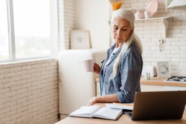 Üst düzey düşünceli kadın mutfakta dizüstü bilgisayarıyla çalışıyor, elinde bir fincan çayla.
