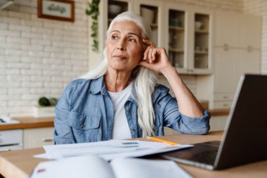 Mutfakta dizüstü bilgisayarıyla çalışan kıdemli gülümseyen kadın.