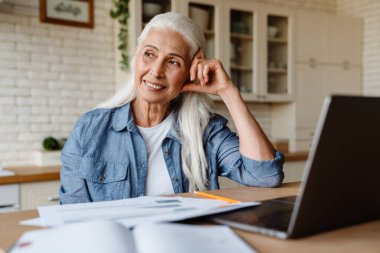 Mutfakta dizüstü bilgisayarıyla çalışan kıdemli gülümseyen kadın.