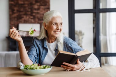 Mutfakta oturmuş salata yerken gülümseyen son sınıf öğrencisi kadın.