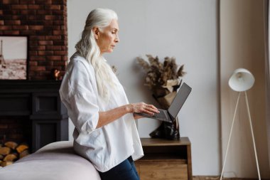 Modern oturma odasında dururken dizüstü bilgisayar kullanan çekici bir yaşlı kadın.