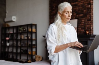 Modern oturma odasında dururken dizüstü bilgisayar kullanan çekici bir yaşlı kadın.