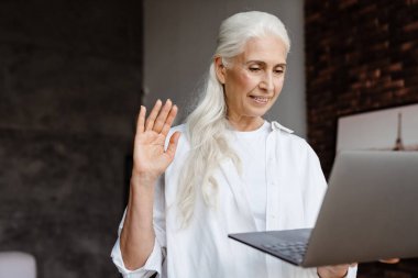 Modern oturma odasında videolu yayında dizüstü bilgisayar kullanan çekici bir yaşlı kadın.