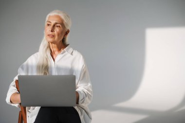 Odaklanmış beyaz saçlı, laptopla çalışan yaşlı kadın gri arka planda izole edilmiş.