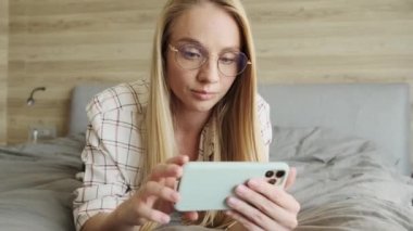 Gözlük takan genç ve güzel bir sarışın kadın evdeki yatak odasında akıllı telefonunu kullanıyor.