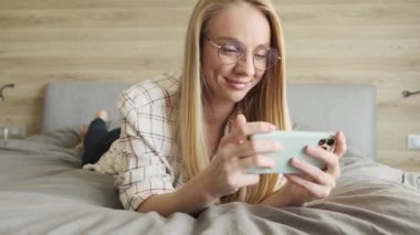 Gözlüklü, gülümseyen sarışın bir kadın, akıllı telefonunu yatak odasında yatay olarak tutarken izliyor.