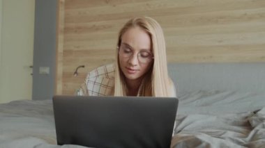 Gözlüklü genç, pozitif sarışın bir kadın evdeki yatak odasında dizüstü bilgisayarını kullanıyor.