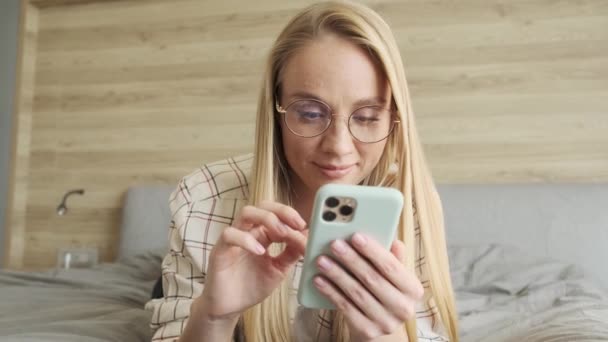 Vakker Ung Blondine Med Briller Bruker Smarttelefonen Sin Soverommet Hjemme – stockvideo