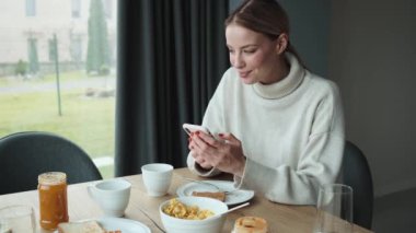 Güzel sarışın kadın kahvaltısını yaparken akıllı telefonunu kullanıyor. Dairelerinde oturuyor.