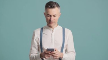 Düşünen bir adam mavi arka plan duvarında izole halde duran akıllı telefonunu kullanıyor.