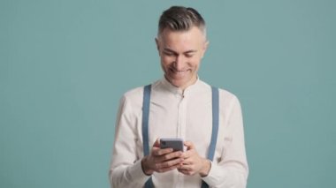 Gülen bir adam mavi arka plan duvarında duran akıllı telefonunu kullanıyor.