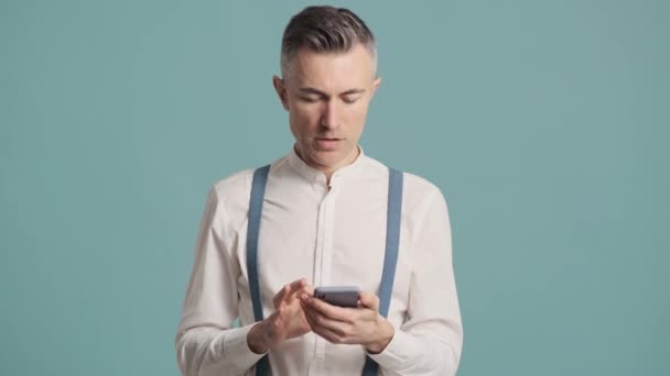 一个吃惊的男人用他的智能手机站在蓝色的背景墙上 — 图库视频影像