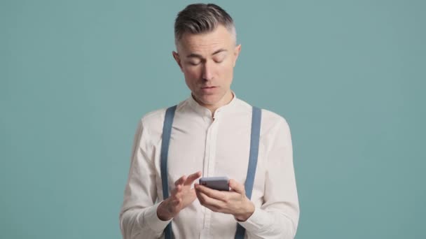 一个快乐的男人站在蓝色的背景墙上 用他的智能手机做着获胜的手势 — 图库视频影像