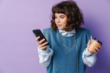 Kafası karışmış güzel bir kız, akıllı telefon kullanıyor ve mor arka planda izole edilmiş kahve içiyor.