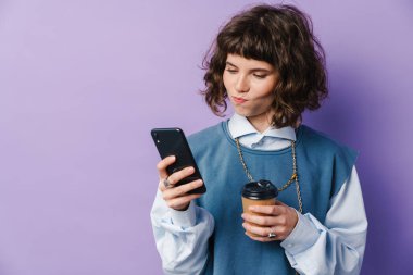 Şaşkın güzel kız akıllı telefon kullanıyor ve mor arka planda izole edilmiş kahve içiyor.