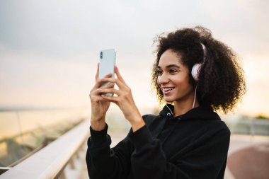 Kulaklıklı neşeli sporcu kadın şehir köprüsünde cep telefonuyla fotoğraf çekiyor.
