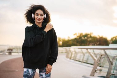 Kendine güvenen genç Afrikalı sporcu kadın köprüde egzersiz yapıyor, kulaklık takıyor.