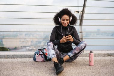 Şehir köprüsünde otururken kulaklıklı neşeli sporcu kadın cep telefonu kullanıyor.