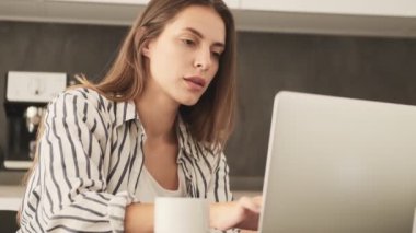 Odaklanmış genç bir kadın evde mutfaktayken dizüstü bilgisayarını kullanıyor.