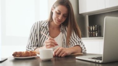 Yoğunlaşmış genç bir kadın evde mutfakta oturmuş kahvaltı ederken bir şeyler yazıyor.