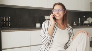 Gözlüklü, gülümseyen genç bir kadın evde mutfakta otururken akıllı telefonuyla konuşuyor.