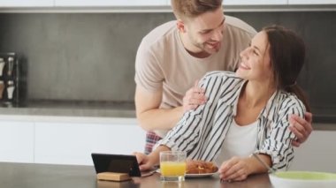 Gülümseyen genç bir çift kadın ve erkek evdeki mutfakta kahvaltı sırasında oturup video izliyorlar.