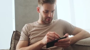 Odaklanmış genç bir adam akıllı telefonunu Stylus 'la birlikte evde koltukta çalışıyor.