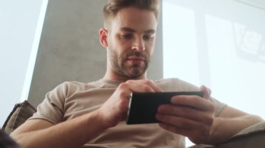 Gülümseyen genç bir adam akıllı telefonunu Stylus 'la evde kanepede otururken kullanıyor.