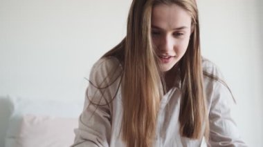 Gülümseyen bir kadın evdeki beyaz yatak odasındaki yatakta otururken dizüstü bilgisayarını kullanıyor.