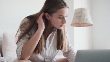 Güzel bir kadın evdeki beyaz yatak odasındaki yatakta otururken dizüstü bilgisayarını kullanıyor.