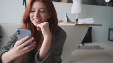 Memnun bir kızıl kadın evde akıllı telefonunu kullanıyor.