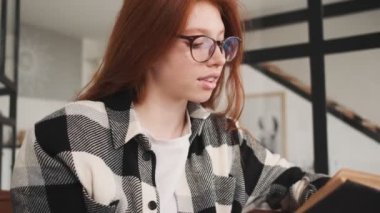 Sakin, gözlüklü, kızıl saçlı bir kadın apartmanda oturmuş kitap okuyor.