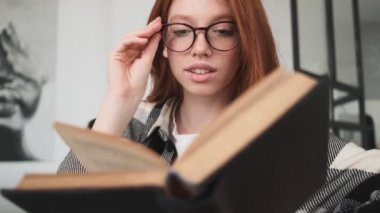 Gözlüklü, pozitif kızıl saçlı bir kadın apartmanda oturmuş kitap okuyor.