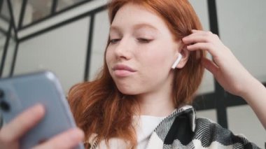 Kulaklık takan kızıl saçlı bir kadının yakın çekim görüntüsü dairesinde oturan akıllı telefonunu kullanıyor.