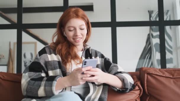 Happy Redhead Woman Earbuds Using Her Smartphone Sitting Sofa Apartments — Stock Video