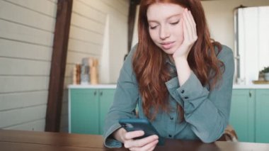 Mutlu, genç, kızıl saçlı bir kadın apartmandaki masada oturan akıllı telefonunu kullanıyor.