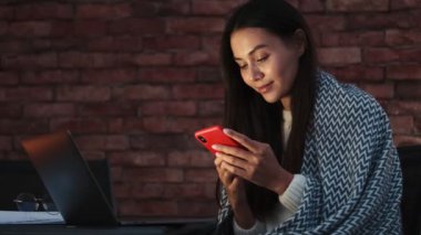 Memnun genç Asyalı bir kadın dairelerin içinde dizüstü bilgisayarıyla çalışırken akıllı telefon kullanıyor.