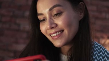 Genç Asyalı mutlu bir kadının yakından görüntüsü terasta oturan akıllı telefonunu kullanıyor.