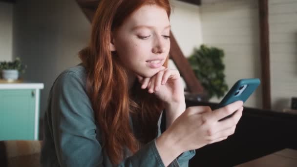 穏やかな若い赤毛の女性は彼女のスマートフォンを使用していますアパートのテーブルに座って — ストック動画