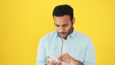 Şaşırmış bir Hintli akıllı telefonunu kullanırken stüdyoda sarı arka planda izole bir şekilde duran baş parmak hareketi gösteriyor.