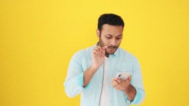Mutlu bir Hintli adam akıllı telefonunu bir tarafı işaret etmek yerine kullanıyor ve sarı arka planda tek başına stüdyoda duruyor.