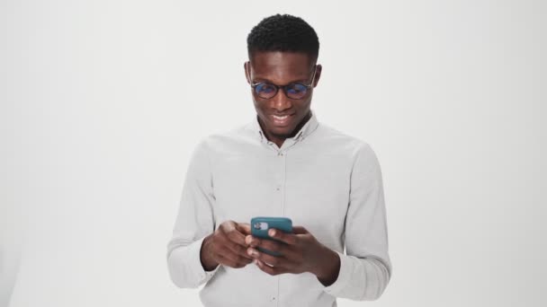 一个戴着眼镜的快乐的非洲裔美国人在工作室里用他的智能手机站在白色背景下做着获胜的手势 — 图库视频影像