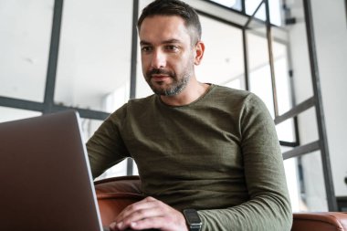 Modern bir ofiste çalışan yakışıklı genç işadamı, dizüstü bilgisayar kullanıyor, kanepede oturuyor.